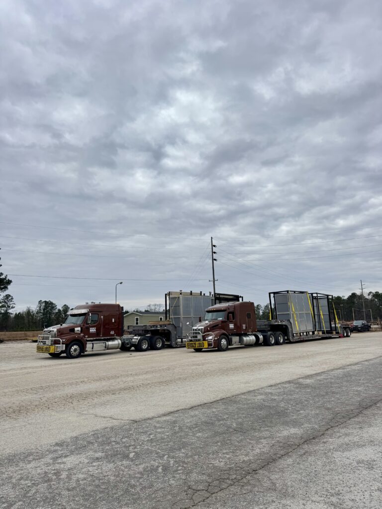 Two semi tractor rigs with lowboy trailers parked at a yard under cloudy sky.