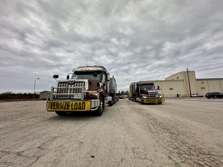 Two heavy-haul trucks with oversized load signs parked in a yard with industrial buildings and cloudy sky in background.