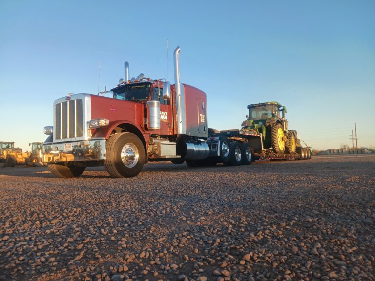Semi tractor with RGN trailer carrying a green John Deere tractor. Road equipment visible in background.