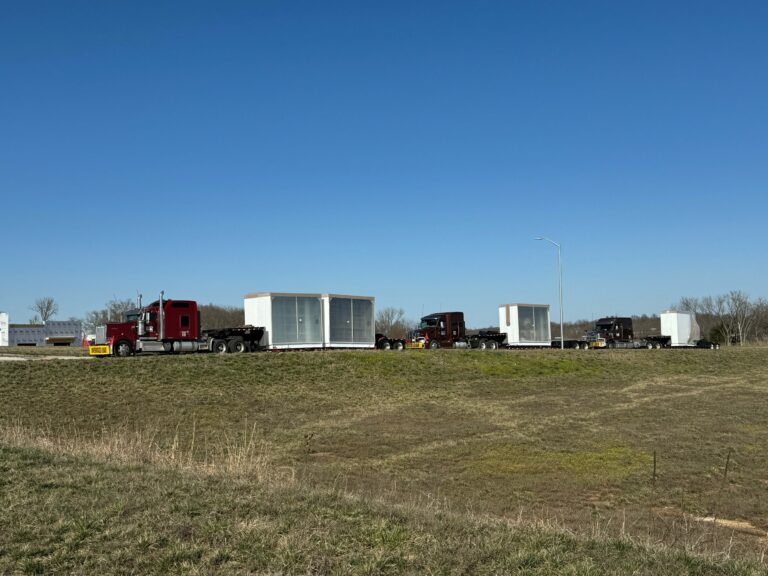 Multiple semi tractors parked in a row with white enclosed trailers in an open yard under clear sky