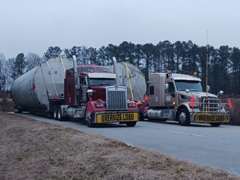Two tractor-trailers parked on roadside with oversized load escort vehicle, marked with OVERSIZE LOAD signs, forested background.