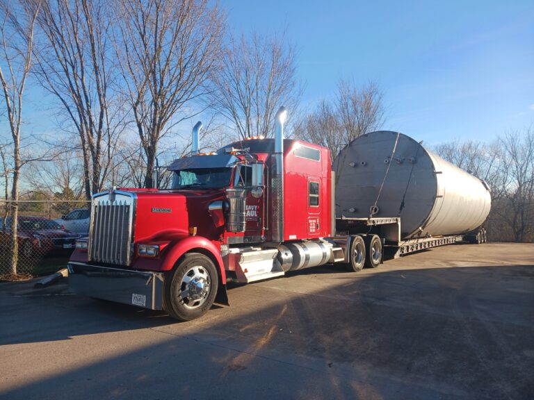 Semi tractor with red cab parked beside bare trees, pulling a large cylindrical silver tank.