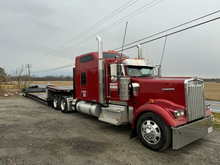 Semi tractor with RGN trailer parked on gravel lot, overcast sky and open fields in background