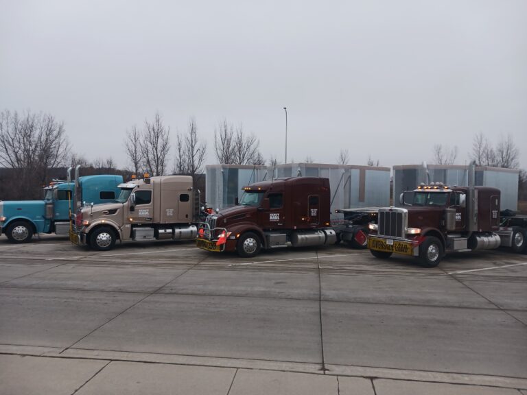 Four semi tractors parked in a row on pavement with bare trees and trailers visible in background.