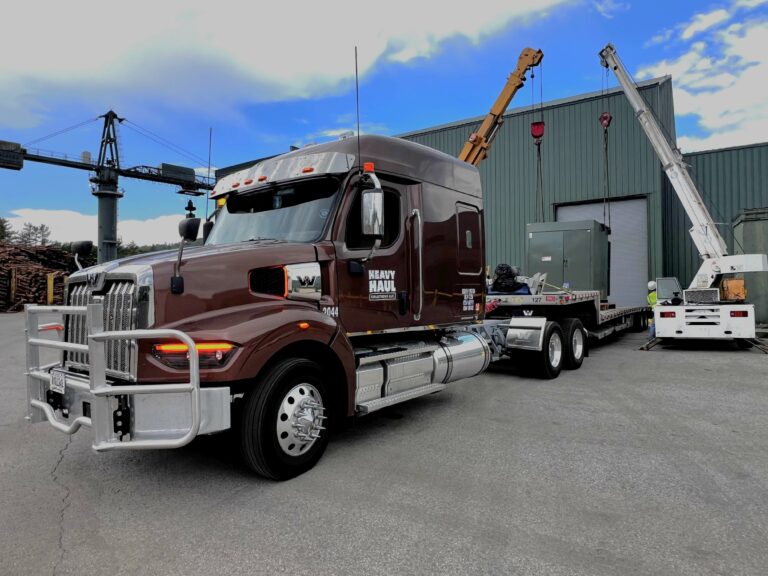 Semi tractor with step deck trailer parked at industrial yard with crane and equipment visible in background.