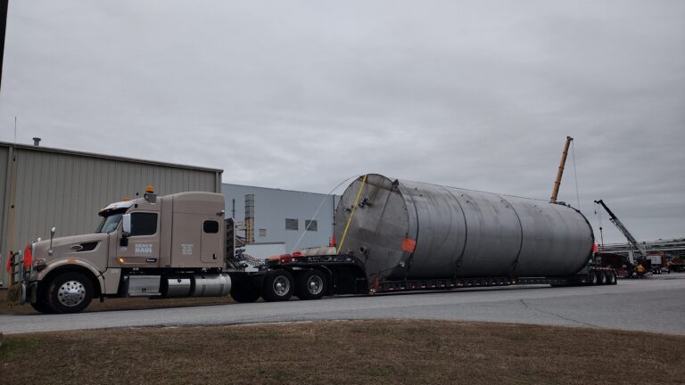 Semi tractor with RGN trailer carrying large cylindrical industrial tank, parked at industrial facility with warehouse building in background.