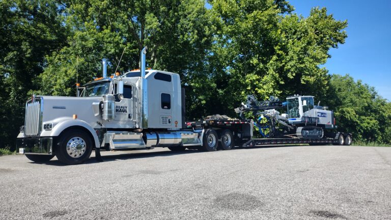Heavy Haul Solutions semi tractor with step deck trailer parked in lot, hauling industrial equipment and machinery, trees in background.