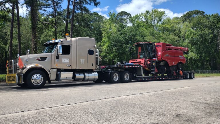 Semi truck with tandem RGN trailer hauling a red combine harvester, parked in a lot with pine trees behind.