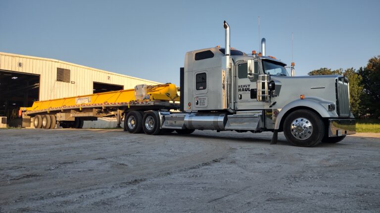Semi tractor with flatbed trailer carrying yellow beam parked in yard with industrial building in background.
