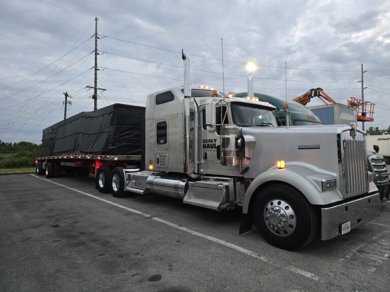 Semi tractor with tall chrome stacks parked next to flatbed trailer loaded with dark cargo material at an industrial facility.