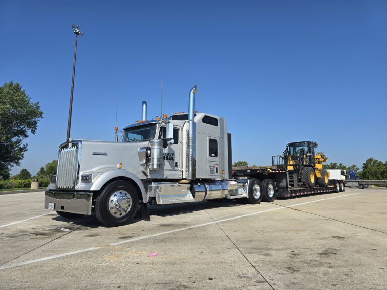 Semi tractor with step deck trailer parked on concrete pad, loaded with yellow wheel loader. Trees and clear blue sky in background.