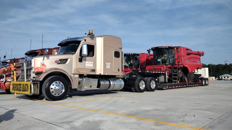 Heavy Haul Solutions semi tractor towing lowboy trailer loaded with large red industrial machinery in a paved yard under clear sky.