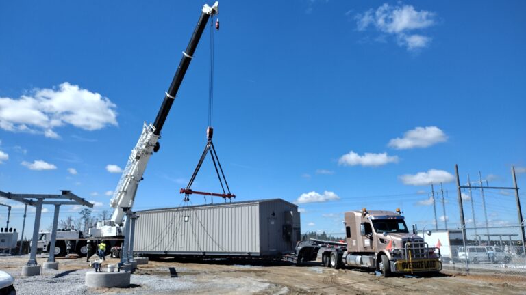 Heavy-duty crane lifting a large industrial module onto an RGN lowboy trailer at an industrial yard under clear skies.