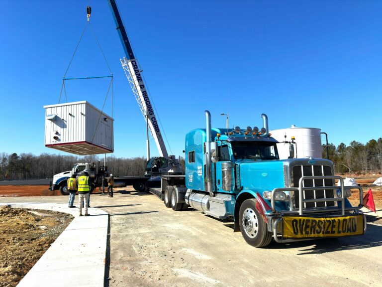 Semi truck with crane lifting modular building unit at construction site with workers in safety gear
