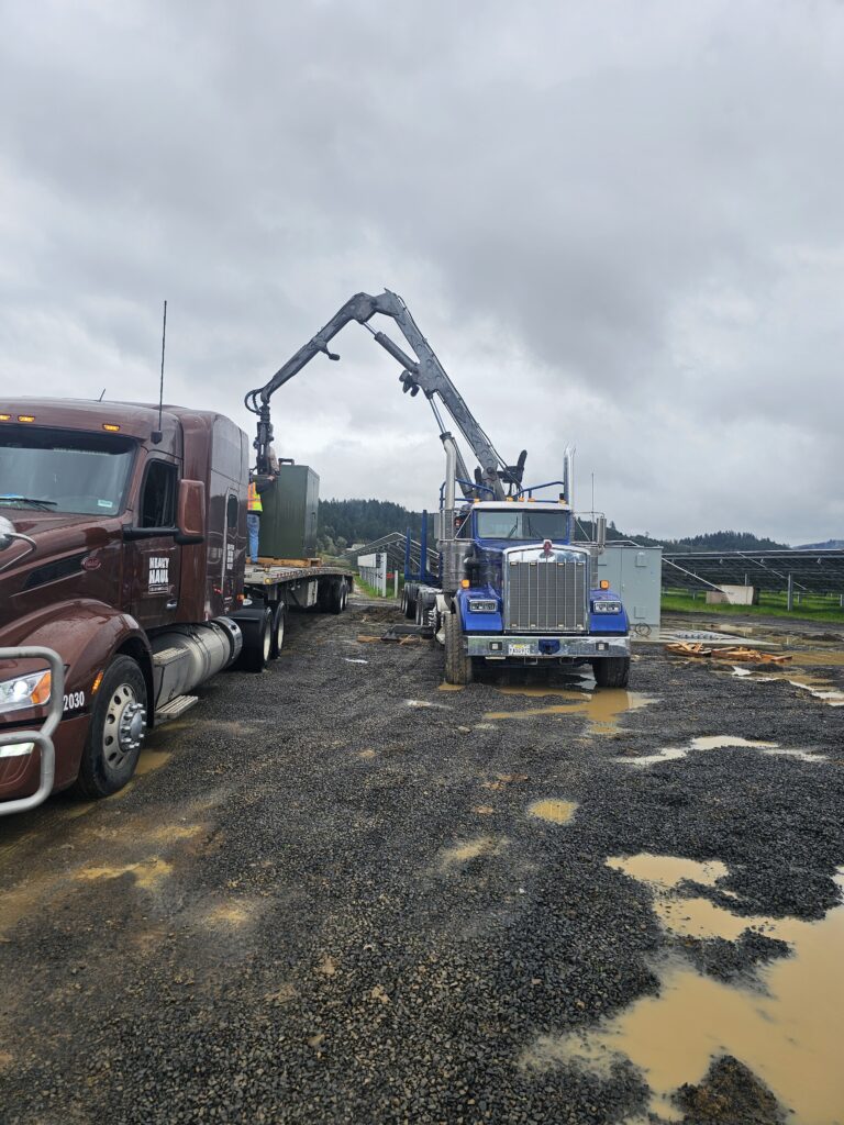 Truck with hydraulic crane arm parked at gravel lot, forested hills in background under overcast sky.