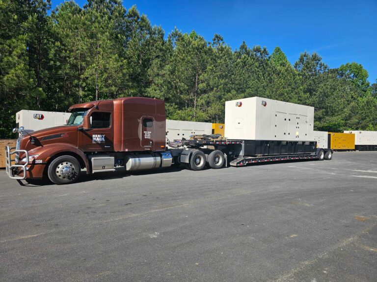 Semi tractor with RGN trailer loaded with large white industrial units and yellow generator, parked in yard with pine trees in background.