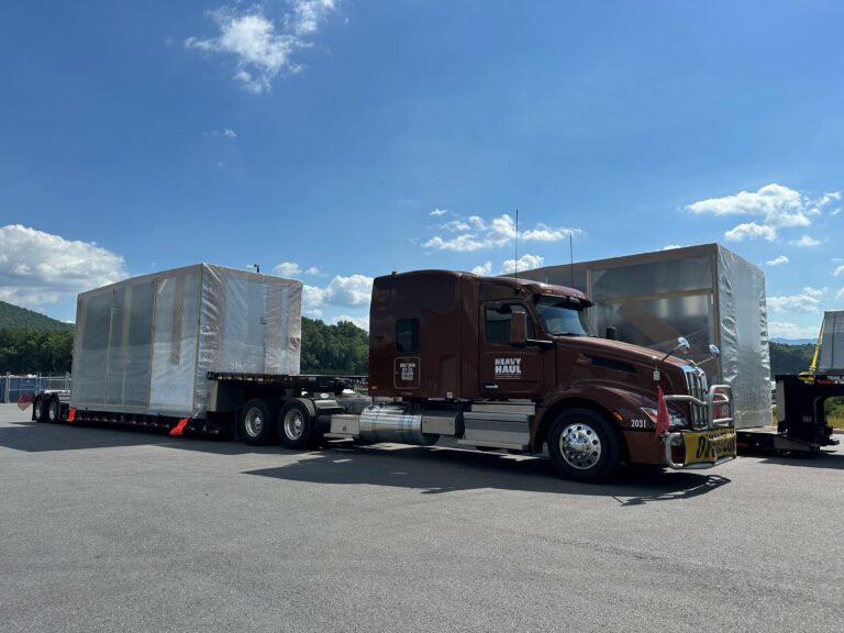 Semi tractor with brown cab towing white enclosed trailers in a truck yard with mountains and blue sky in background.