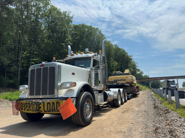 Semi tractor with orange warning lights parked on dirt road alongside equipment trailer; construction site and highway overpass visible in background.