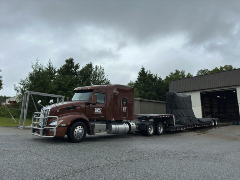 Heavy Haul Solutions semi truck with RGN trailer carrying metal frame structure, parked at industrial yard with buildings and trees in background.