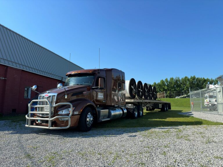 Semi tractor with flatbed trailer carrying large industrial wheels parked at yard near metal building