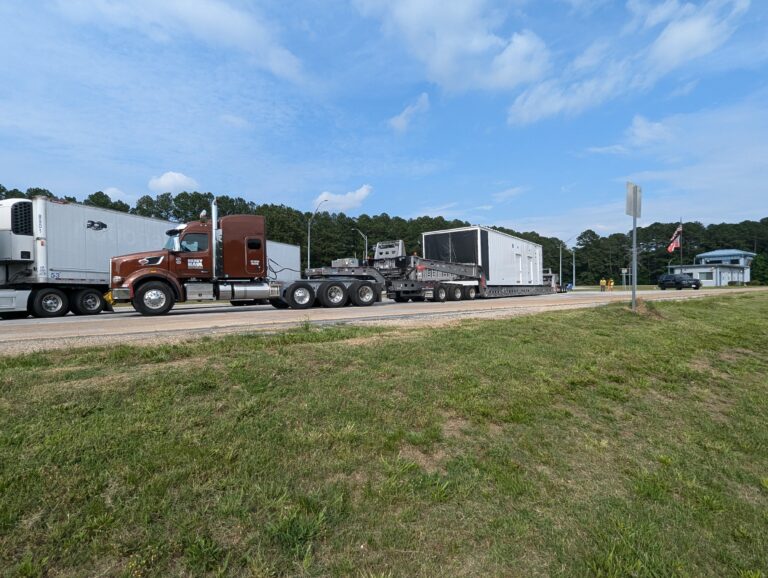 Semi tractor with brown cab pulling multiple trailers loaded with large industrial modules, parked at a facility with grass foreground and trees in background.