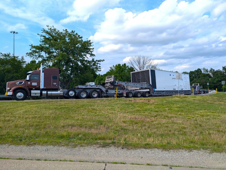 Semi tractor with burgundy cab parked on grass next to step deck trailer carrying large white industrial module.