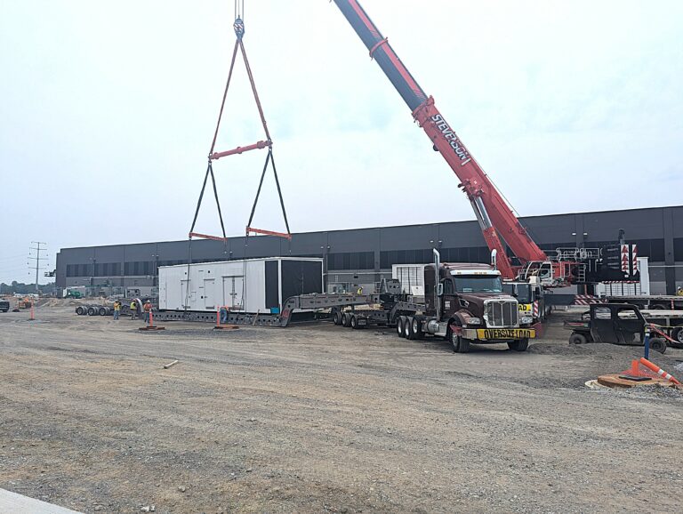 Heavy-haul truck with crane positioned next to a white industrial module suspended by rigging cables in a yard facility.