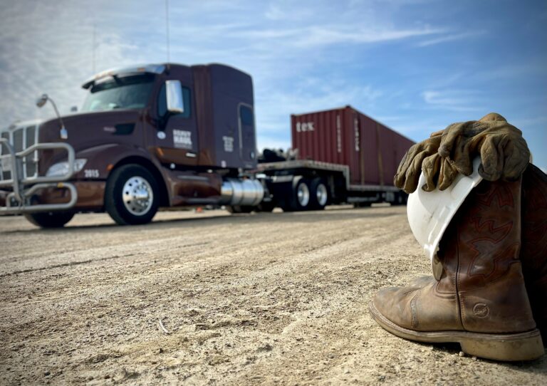 Cowboy boots with white socks rest on gravel with a heavy-haul semi tractor and container trailer parked in the background.