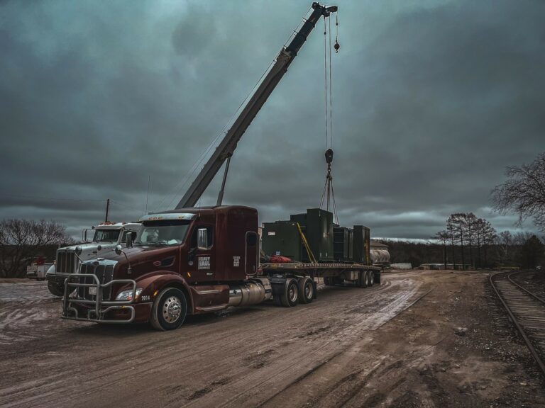 Semi tractor with boom crane parked at industrial yard, flatbed trailer with large enclosed cargo unit suspended by crane cables.