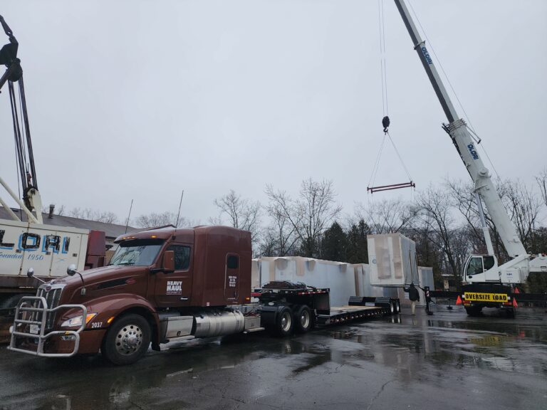 Semi tractor with RGN trailer carrying white industrial modules, with crane lifting equipment overhead in wet yard.