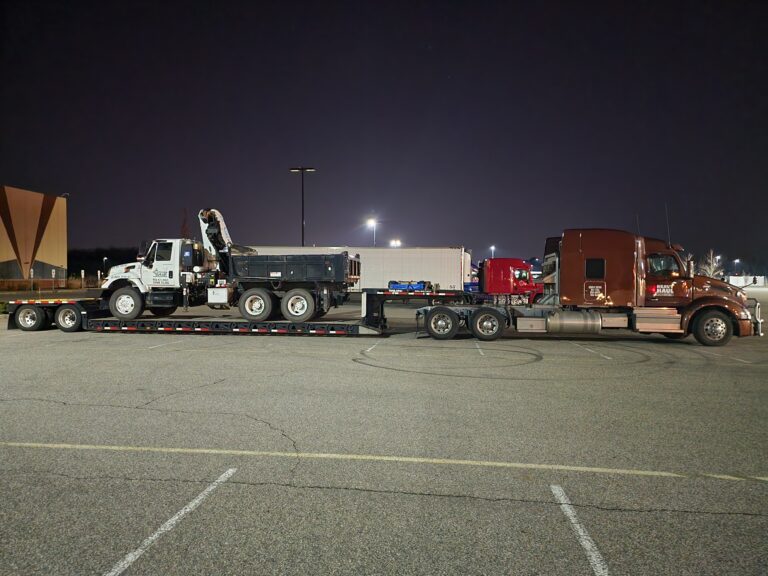 Lowboy trailer with heavy equipment parked in a nighttime yard alongside a dump truck and tractor-trailer.