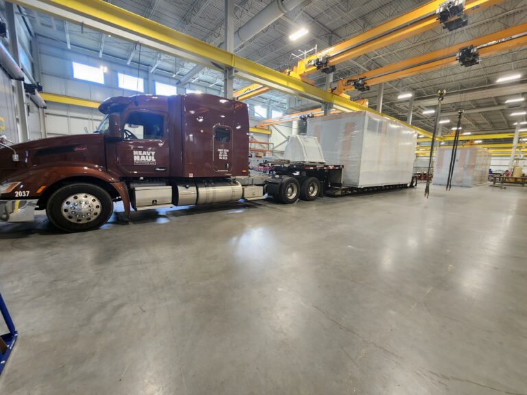Semi tractor with Heavy Haul Solutions branding parked in warehouse with enclosed trailer and yellow overhead cranes visible.