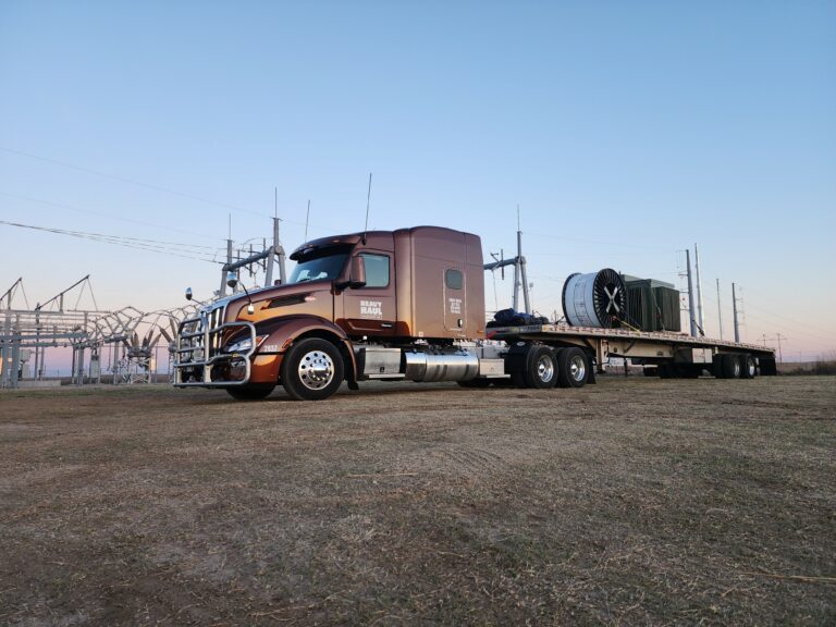 Semi tractor with flatbed trailer carrying large industrial spools parked in yard with industrial structures visible in background.