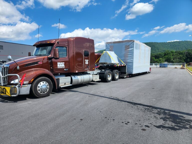 Semi tractor with white step deck trailer parked in industrial yard with forested hills in background