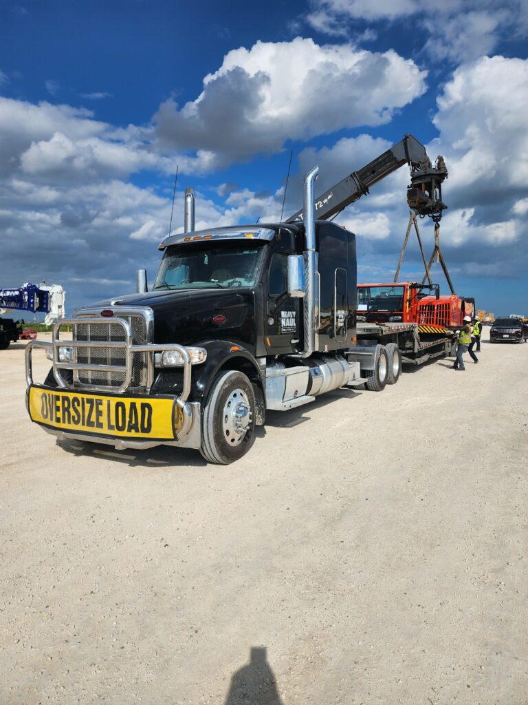 Semi tractor with crane boom parked on dirt lot, oversized load warning sign visible, support vehicle nearby under blue sky.