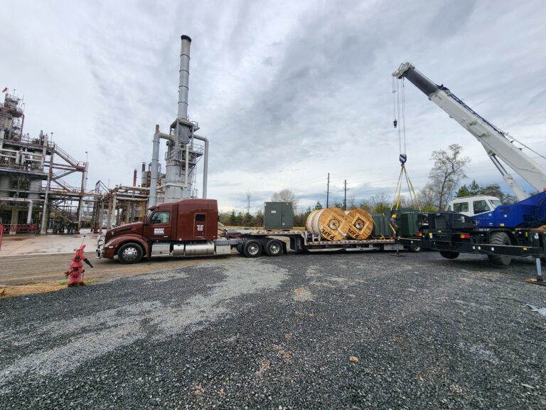 Semi truck with stepdeck trailer carrying large coils of material at an industrial facility with crane and equipment visible.