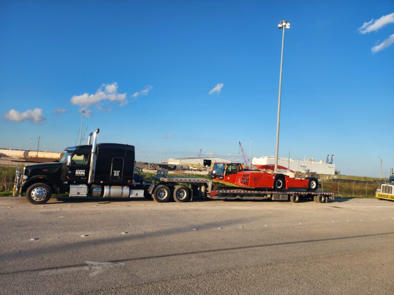 Semi tractor with stepdeck trailer carrying red industrial machinery, parked at a terminal with industrial facilities in background.