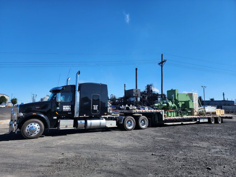 Semi tractor with stepdeck trailer carrying large industrial machinery and equipment parked at a yard under clear blue sky