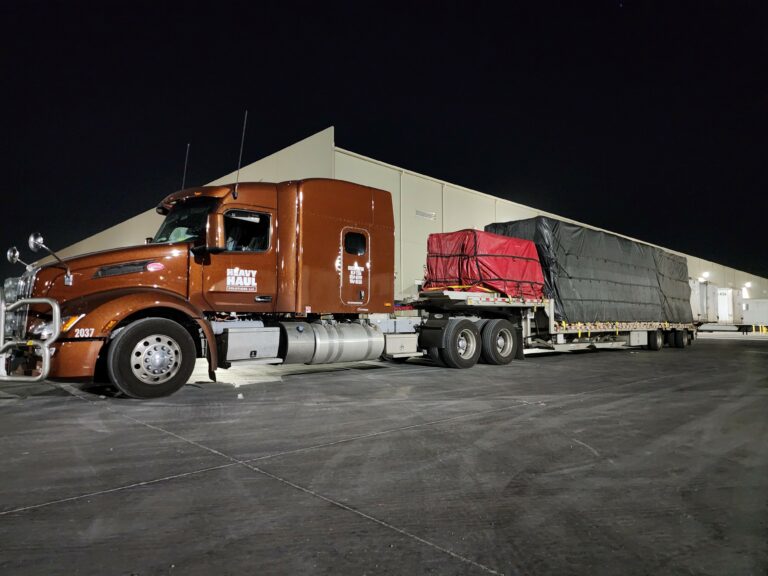 Semi tractor with brown cab and stepdeck trailer carrying red and black tarped cargo, parked at night in front of warehouse with tent structure.