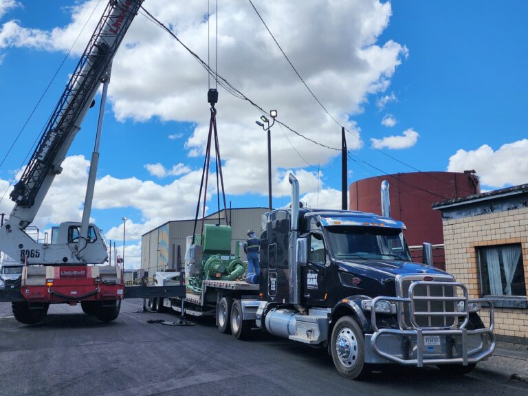 Crane truck with boom hoist positioned near a stepdeck trailer carrying industrial equipment in a yard with buildings and clear sky.