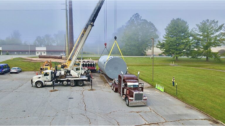 Truck crane positioned beside maroon semi truck with large cylindrical tank suspended by lifting cables over rural industrial facility.