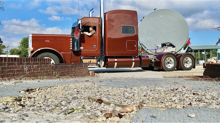 Semi tractor with enclosed sleeper cab parked next to RGN lowboy trailer carrying large satellite dish at yard facility.