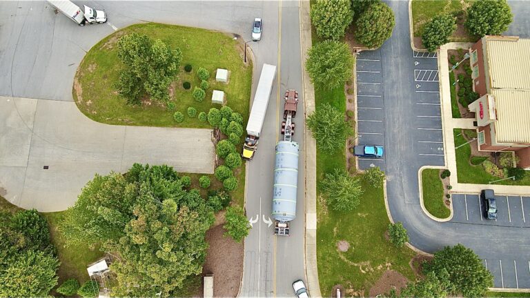 Aerial view of a tanker trailer parked on a residential street next to a grassy lot and parking areas with trees and buildings visible.