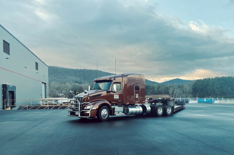 Heavy Haul Solutions semi tractor with RGN trailer parked at a yard with industrial building and mountains in background.