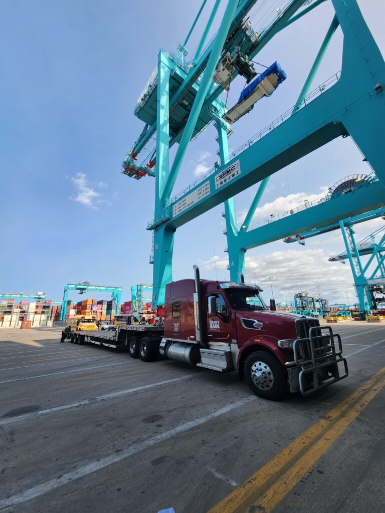 Semi truck with stepdeck trailer parked beneath turquoise shipping container cranes at a port terminal with stacked cargo containers in background.