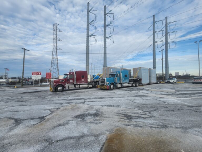 Semi tractor and RGN trailer with large industrial modules parked in industrial yard with electrical transmission towers in background.