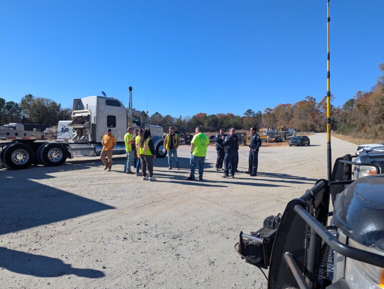 Team members in safety vests gathered in a dirt yard next to a parked semi tractor and various vehicles and equipment.