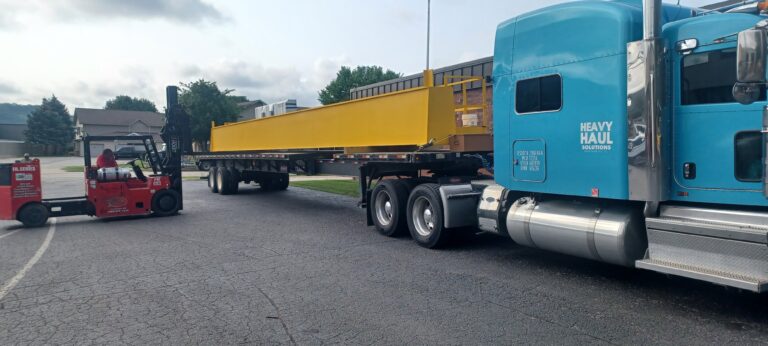 Trailer with yellow bean parked hauled by turquoise Heavy Haul Solutions tractor and red forklift in yard.