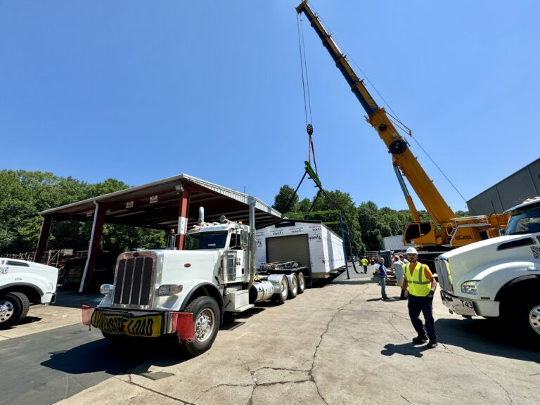 Semi tractor with enclosed trailer parked at yard. Yellow mobile crane with extended boom and workers in safety vests nearby. Metal building and other vehicles in background under clear sky.