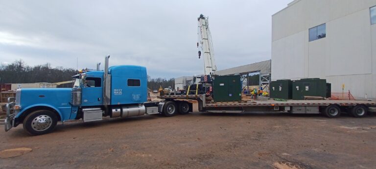Heavy Haul semi truck with stepdeck trailer hauling green industrial transformers near a building with a crane.